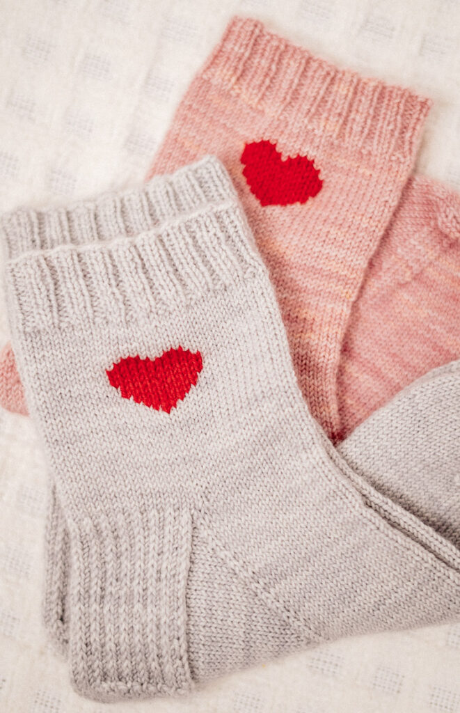 Stack of hand-knit heart socks in soft pink yarn – a perfect handmade gift for Valentine’s Day.