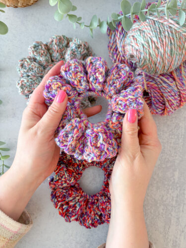 Hand holding a single finished DIY scrunchie made with I Love This Yarn Print, with other scrunchies blurred in the background.