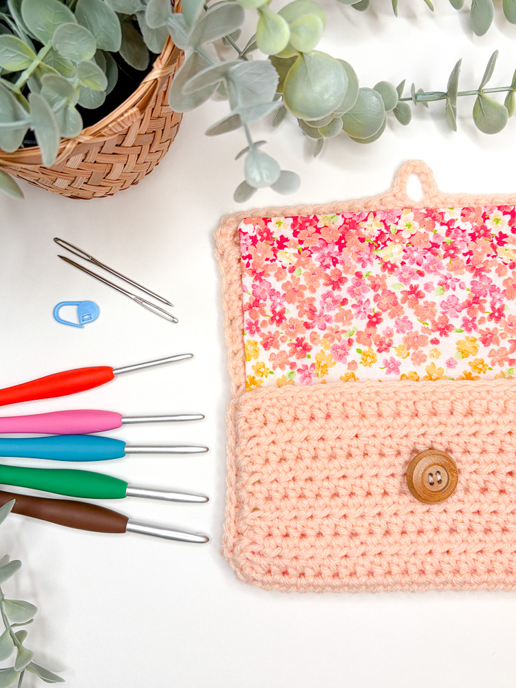 peach crochet hook pouch laying open on a white background with crochet hooks, yarn needles, and a stitch marker next to it